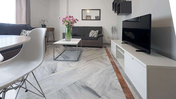 Un salon moderne avec un canapé gris, une table basse blanche, un téléviseur posé sur un meuble blanc et un vase rempli de fleurs.