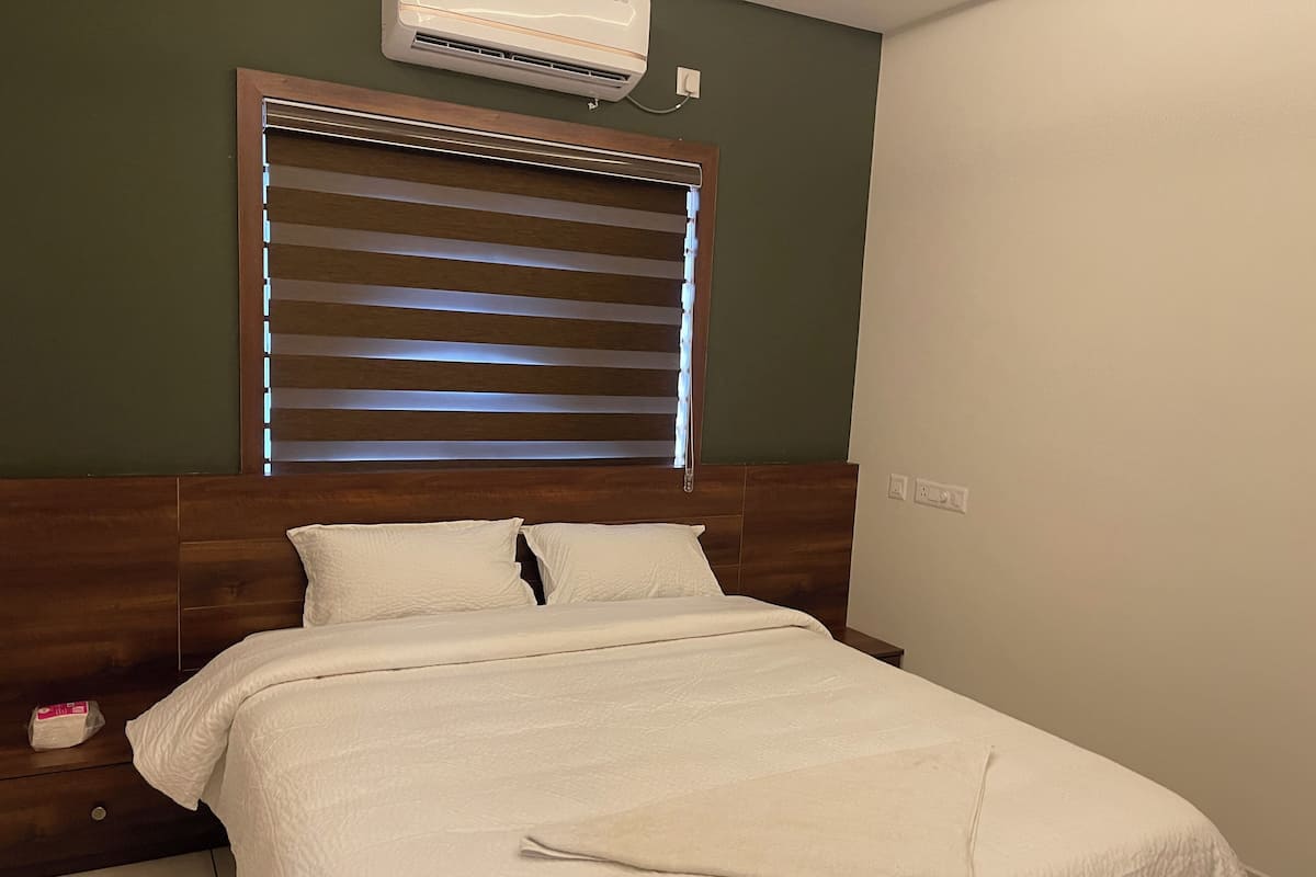 A neatly made bed with a wooden headboard, a window with blinds, and a wall-mounted air conditioner.
