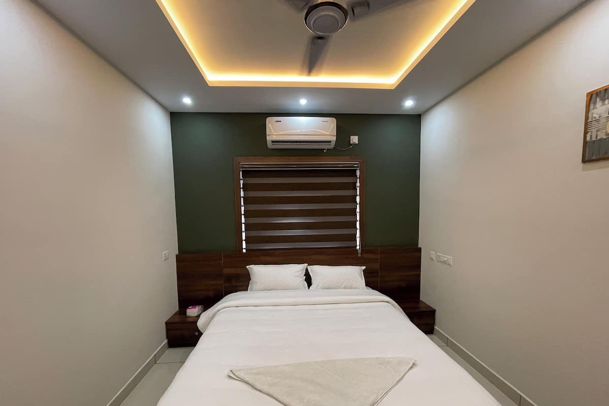 A bedroom with a bed, a ceiling fan, recessed lighting, an air conditioner, a window with blinds, and a nightstand with a lamp.