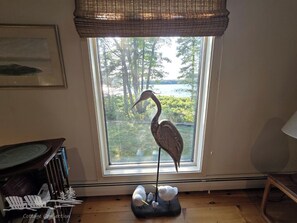 A room with a large window, a framed painting on the wall, a wooden table, and a decorative bird sculpture.