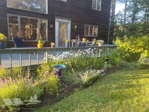 Una casa de madera con porche cubierto, plantas en macetas y un jardín con diversas flores y arbustos.