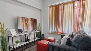 A modern living room with a grey sofa, red ottomans, a TV on a black stand, and a window with sheer curtains. - Updated Loft in a Convenient Area! (Wheat Ridge)