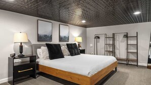 A modern bedroom with a wooden bed, two nightstands with lamps, a ceiling with a geometric pattern, and two framed pictures on the wall. - Industrial-Chic Loft with King Bed Close to Shops! (Wheat Ridge)