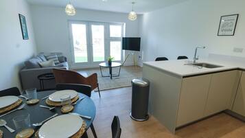A modern kitchen with a dining area, a sofa, and a TV in the background.