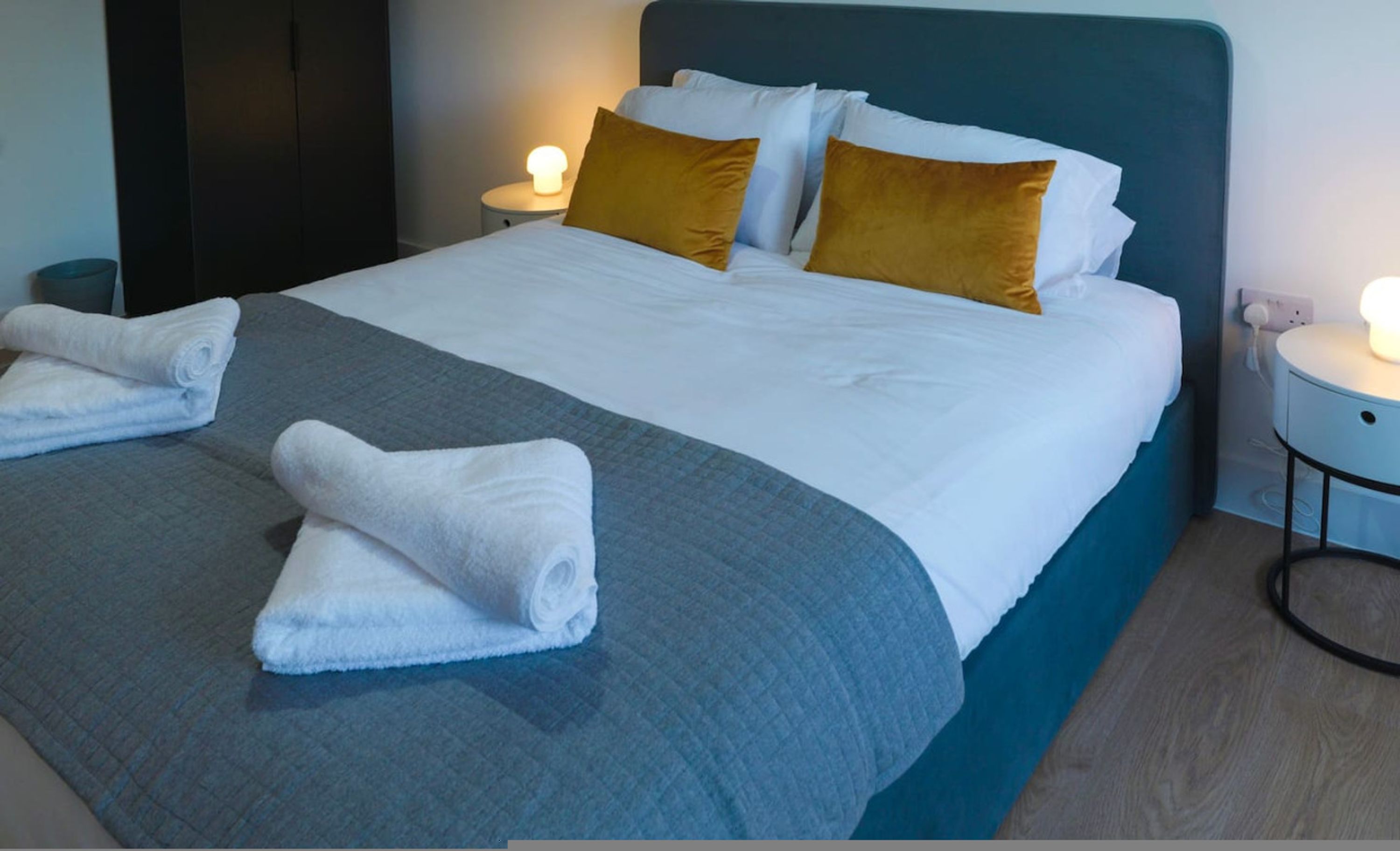 A neatly made bed with white linens, a grey blanket, and two folded towels. There are two bedside tables with lamps, one on each side of the bed.