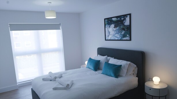 A modern hotel room with a large bed, a bedside table with a lamp, a window with blinds, and a painting on the wall.