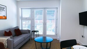 A living room with a gray sofa, a round blue table, a small black table, a black chair, and a dining table with a glass and plate.