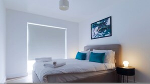 Una camera da letto moderna con una grande finestra, un letto con biancheria da letto bianca e cuscini color acquamarina, un quadro astratto incorniciato e una lampada da comodino.