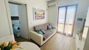 A modern living room with a beige sofa, a painting of a sunset, and a sliding glass door leading to a balcony. - Apartment in beachfront residence (Alghero)