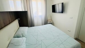 A hotel room with a bed, a TV mounted on the wall, a chair, and a window with curtains. - Apartment in beachfront residence (Alghero)