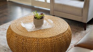 Un pouf rond en fibres tressées, surmonté d’un plateau en verre transparent et agrémenté d’une petite plante en pot, est placé sur un tapis à motifs dans une pièce où se trouvent un canapé et une fenêtre équipée de stores.