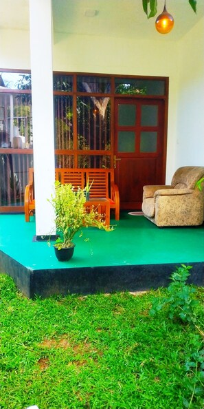 Un porche con piso verde, dos sillas de madera, una planta en maceta y una puerta de madera con paneles de vidrio.