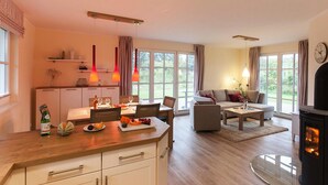 A modern living room with a fireplace, a dining area with a table and chairs, and a kitchen with a wooden countertop. - Ferienhaus, Fuhlendorf (Fuhlendorf)