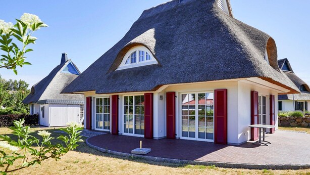 A thatched-roof house with a white exterior and red shutters, featuring a large window and a small patio area. - Ferienhaus, Fuhlendorf (Fuhlendorf)