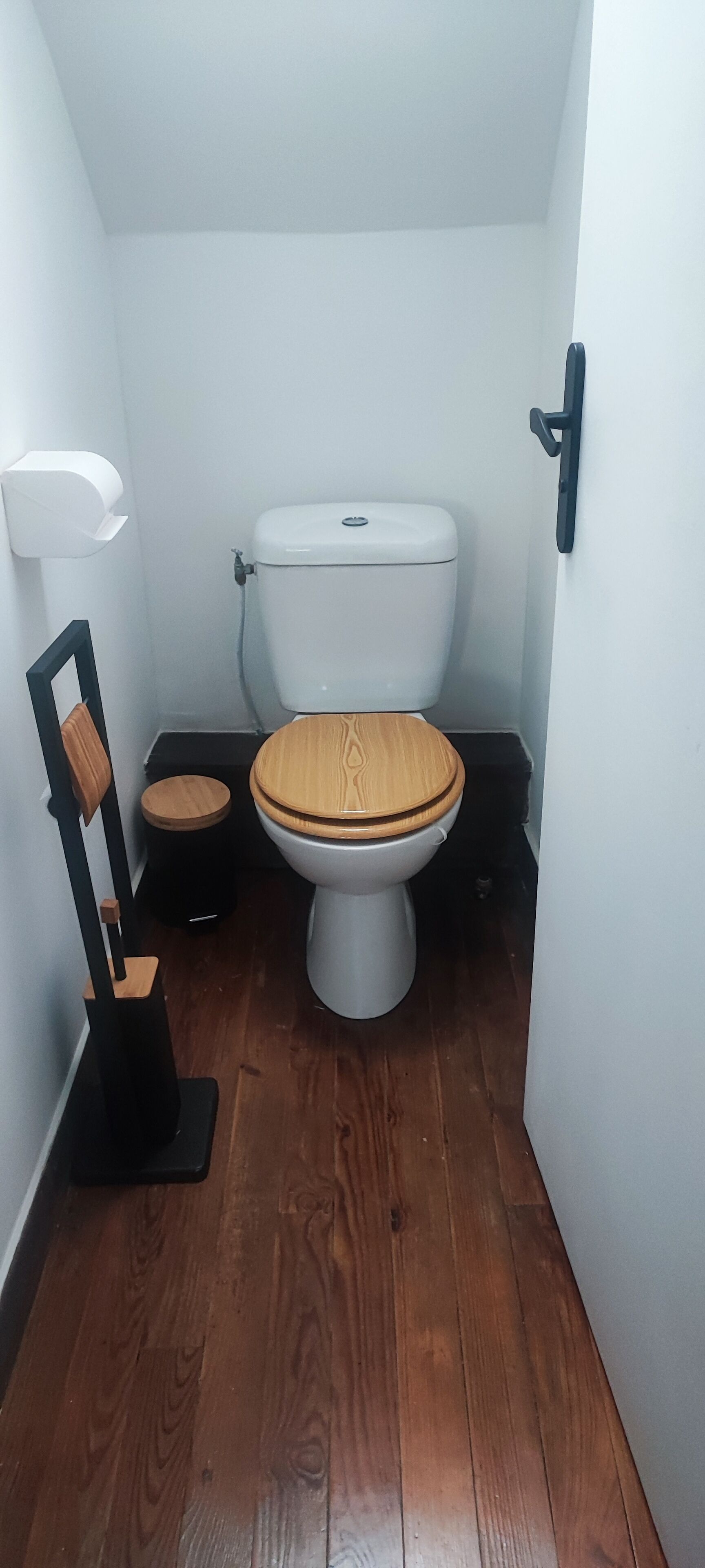 Un bagno piccolo e stretto con un water di legno, un cestino nero e un portasciugamani bianco a parete.