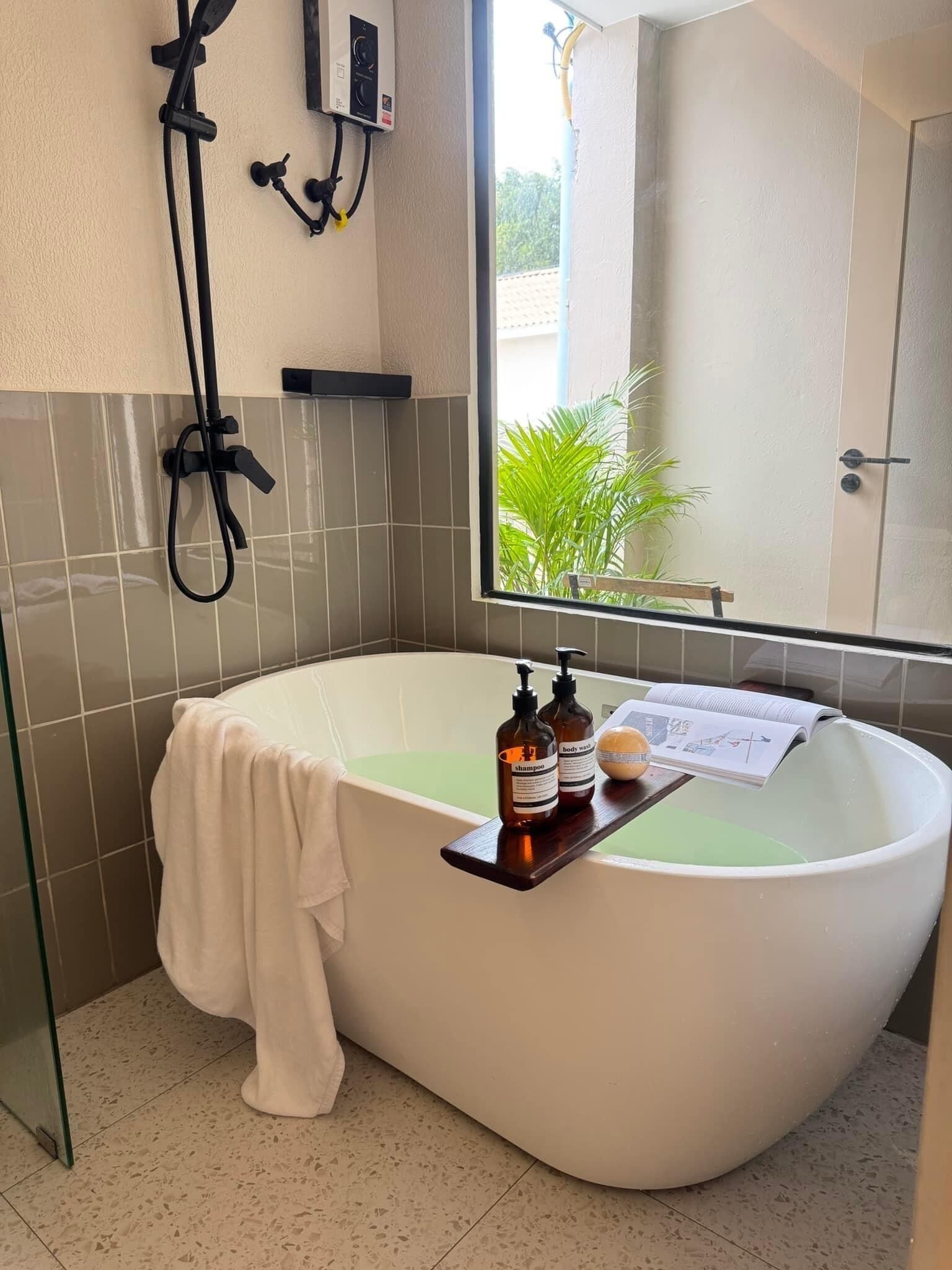 Un baño moderno con una bañera independiente, un perchero, un ventanal con vistas a la vegetación y un estante con artículos de aseo.