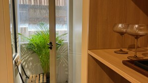 A hotel room with a wooden door, a small seating area with a bench and potted plants, a glass door leading to a balcony, and a shelf with a lamp and a handbag. - Urban Room Samed (Rayong)