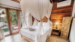 A bedroom with a canopy bed, wooden flooring, a nightstand with a lamp, and a view of a swimming pool through glass doors. - Ganesha Villa (Ubud)