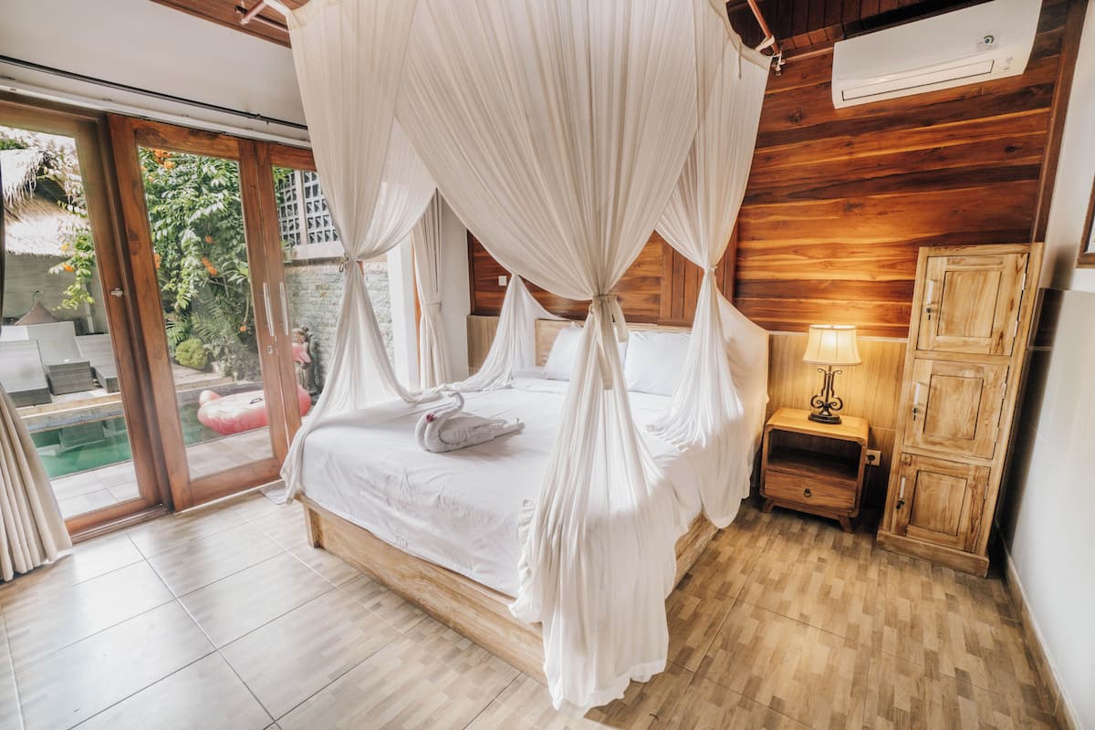 Un dormitorio con cama a dosel, suelo de madera, una mesita de noche con lámpara y vista a una piscina a través de puertas de cristal.