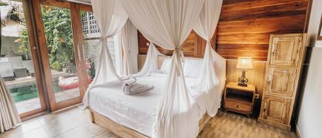 Un dormitorio con cama a dosel, suelo de madera, una mesita de noche con lámpara y vista a una piscina a través de puertas de cristal.