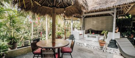 An outdoor patio with a round wooden table and chairs, surrounded by potted plants and a thatched umbrella.