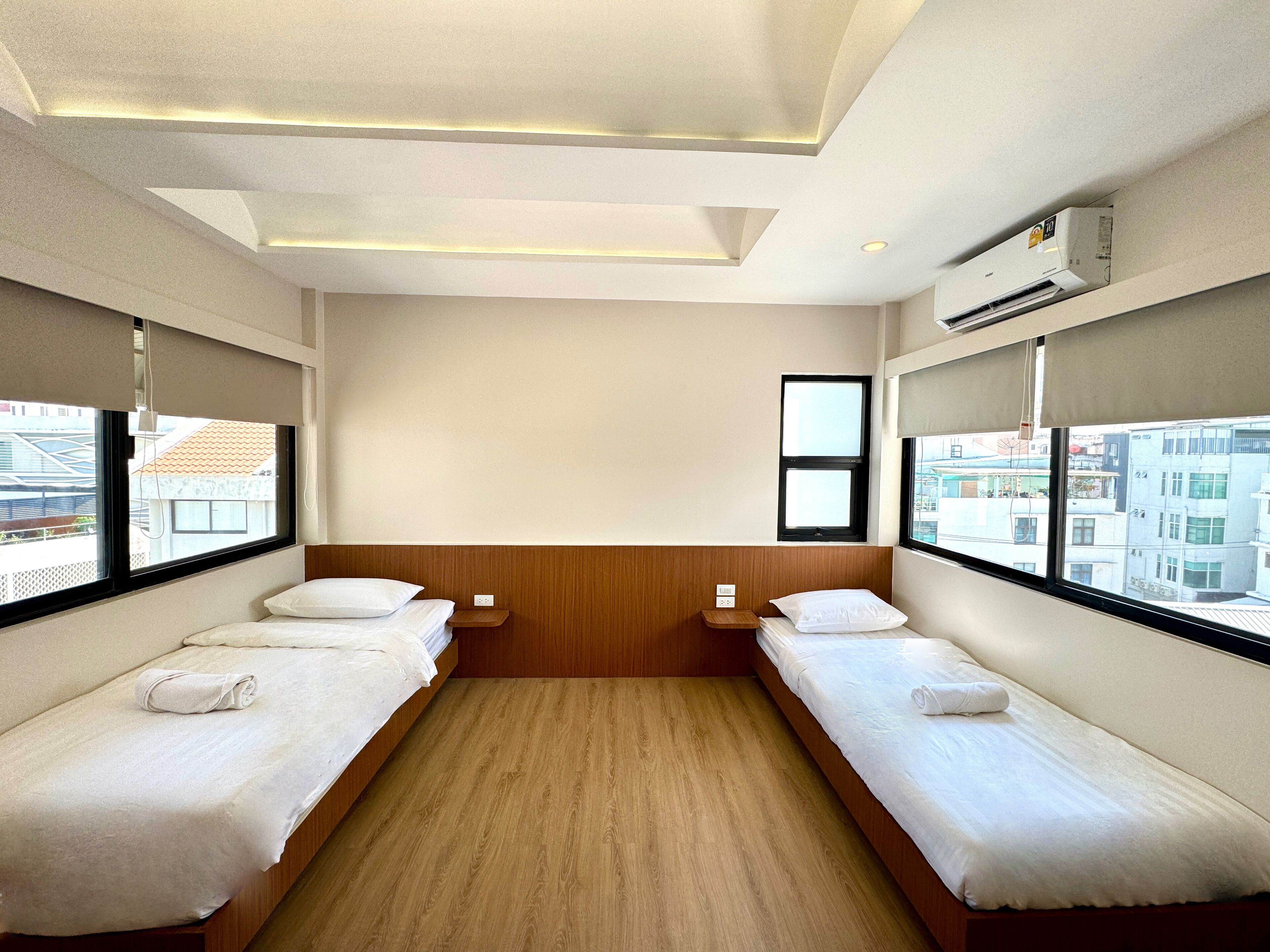 A hotel room with two single beds, a wooden headboard, a small bedside table, a window with a view of buildings, and an air conditioning unit.