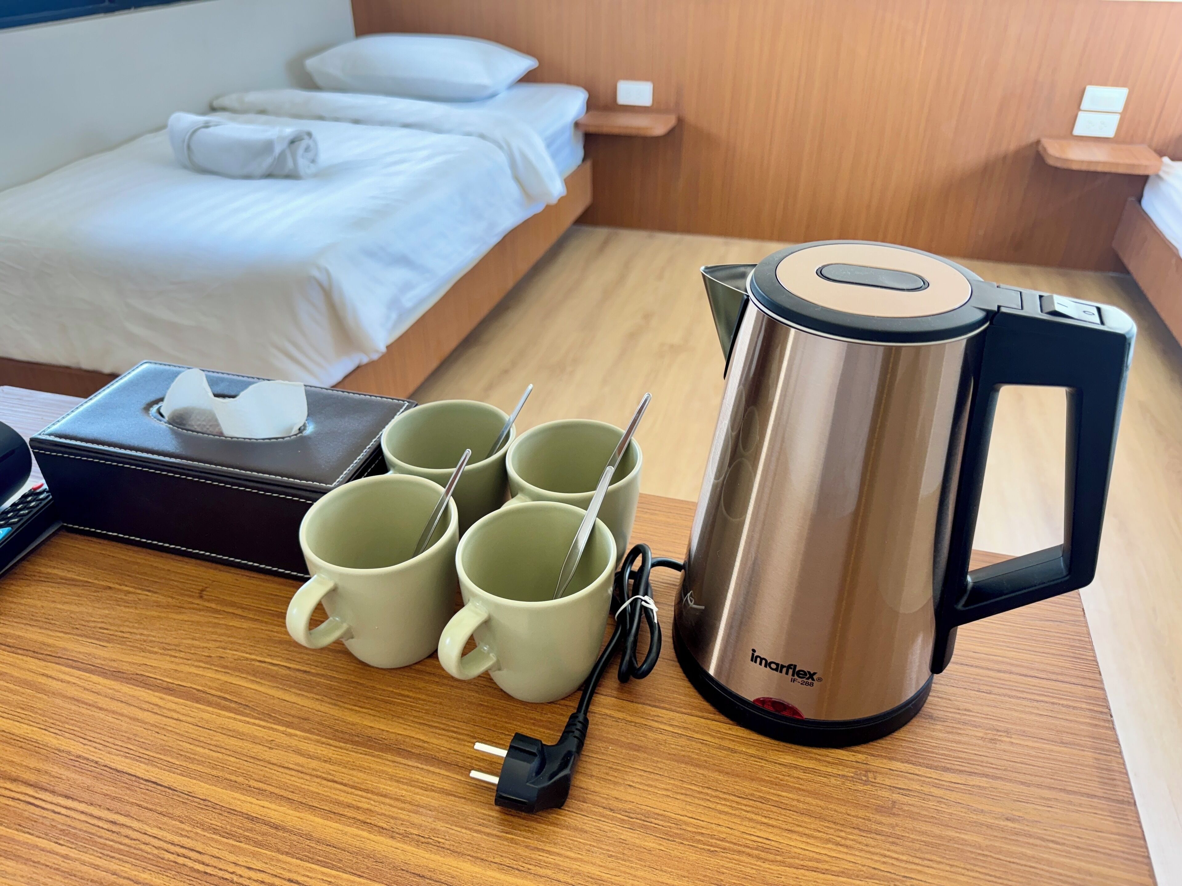 Quarto de hotel com uma cama bem arrumada, uma mesa de cabeceira com caixa de lenços, chaleira e quatro canecas.