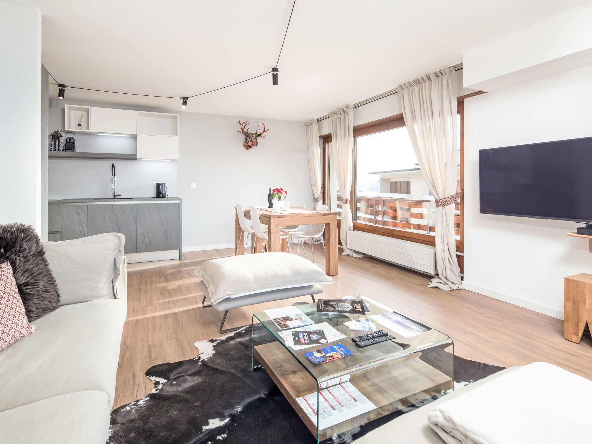 Um apartamento moderno com sala de estar, área de jantar e cozinha compacta. Possui varanda com vista, televisão de tela plana e uma mesa de centro de vidro com revistas e controles remotos.
