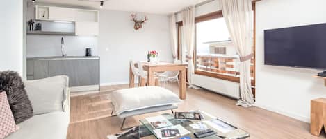 A modern apartment with a living room, dining area, and kitchenette. There is a balcony with a view, a flat-screen TV, and a glass coffee table with magazines and remote controls.