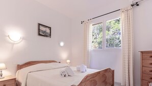 Un dormitorio ordenado con cama de madera, lámparas de noche, una mesita auxiliar pequeña, una ventana con cortinas y un cuadro colgado en la pared.