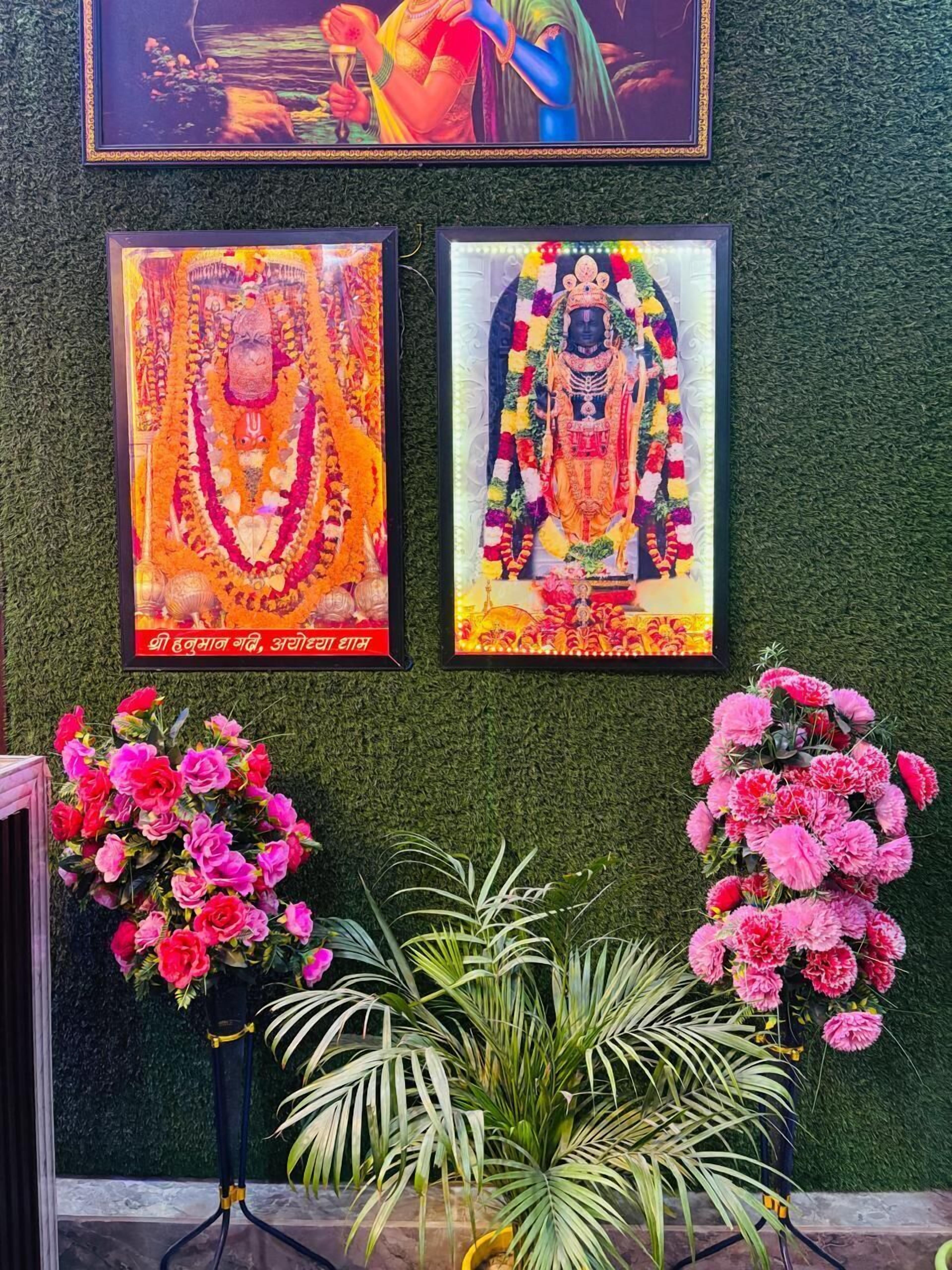 A wall with three framed religious paintings and two potted plants with pink flowers.