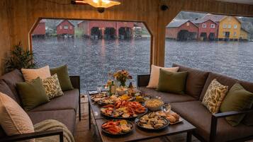 A cozy room with a wooden interior, a dining table set with seafood, and a view of a flooded village through large windows.