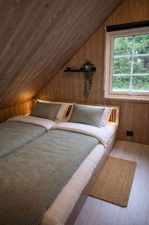 Ein Schlafzimmer mit holzverkleideter Schräge, einem Bett mit gestreifter Bettwäsche, einem Fenster mit Blick auf Grünflächen und einem kleinen Regal mit Dekoration.