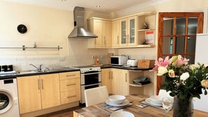 A modern kitchen with wooden cabinets, a stainless steel range hood, a built-in oven, a microwave, and a sink. There is a dining table set with plates, bowls, and a vase of flowers. - Porthilly by Interhome (Mevagissey)