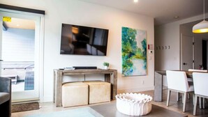 Una sala moderna con un televisor de pantalla plana, un cuadro, un comedor con mesa y sillas, y un rincón de estar acogedor con otomanas.