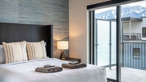 Una habitación de hotel moderna con una cama grande, una mesita de noche con lámpara y vistas a montañas nevadas a través de un ventanal corredero.