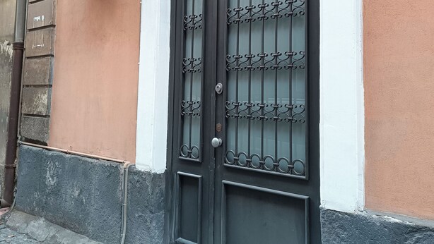 A black metal gate with decorative patterns, attached to a building with a peach-colored wall and a white trim. - Central House Apartment (Catania)