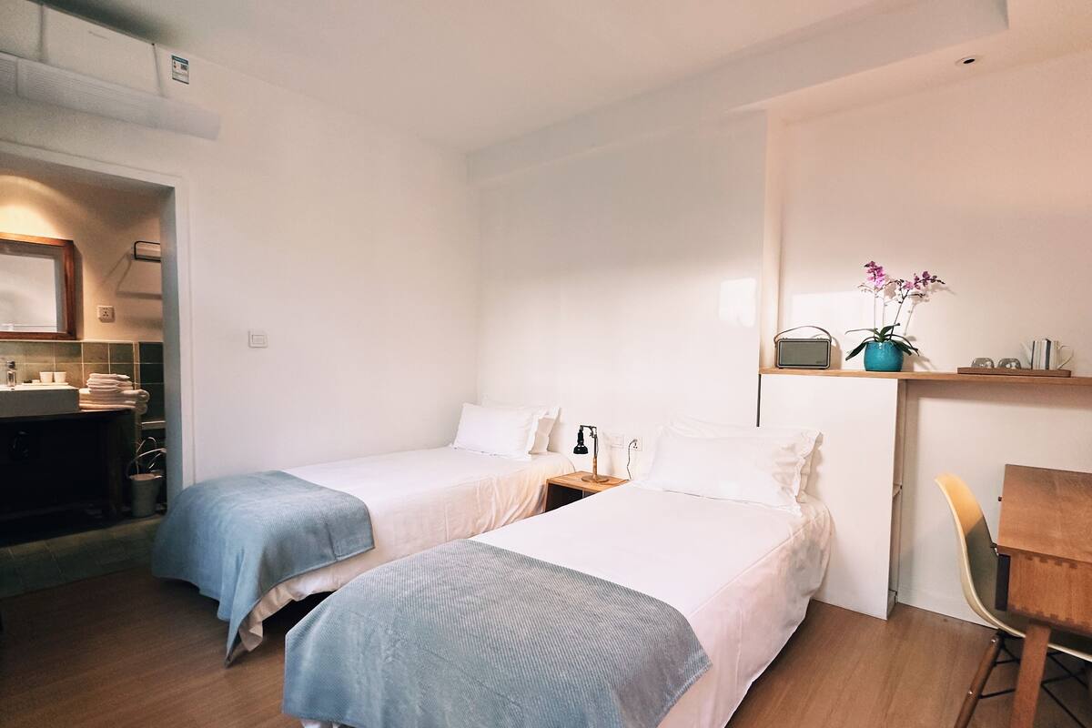 A hotel room with two beds, a wooden desk, a chair, a radio, and a small shelf with a plant.