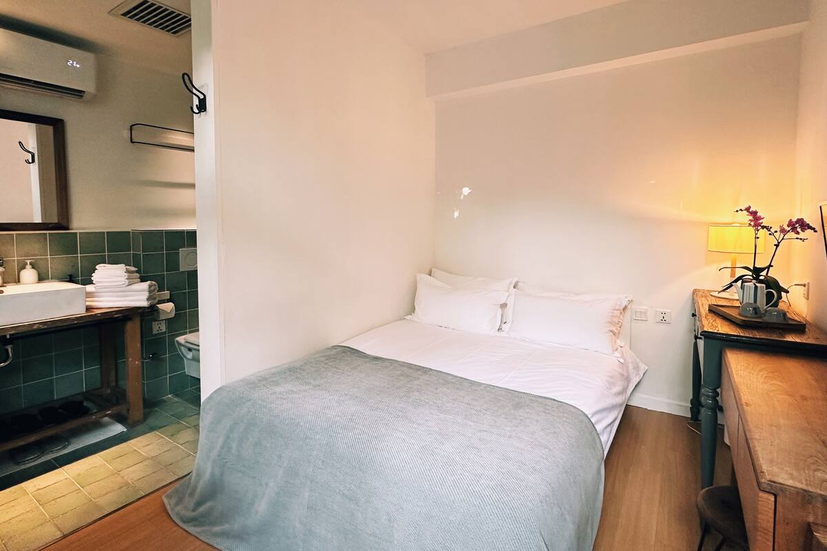 A hotel room with a bed, a bedside table with a lamp and flowers, a bathroom with a sink and towel stack, and a wooden floor.