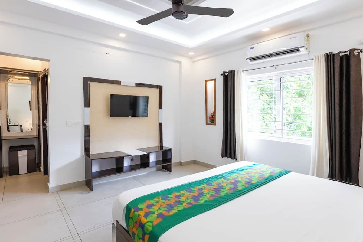 A hotel room with a bed, a TV mounted on the wall, a ceiling fan, an air conditioning unit, and a window with curtains.