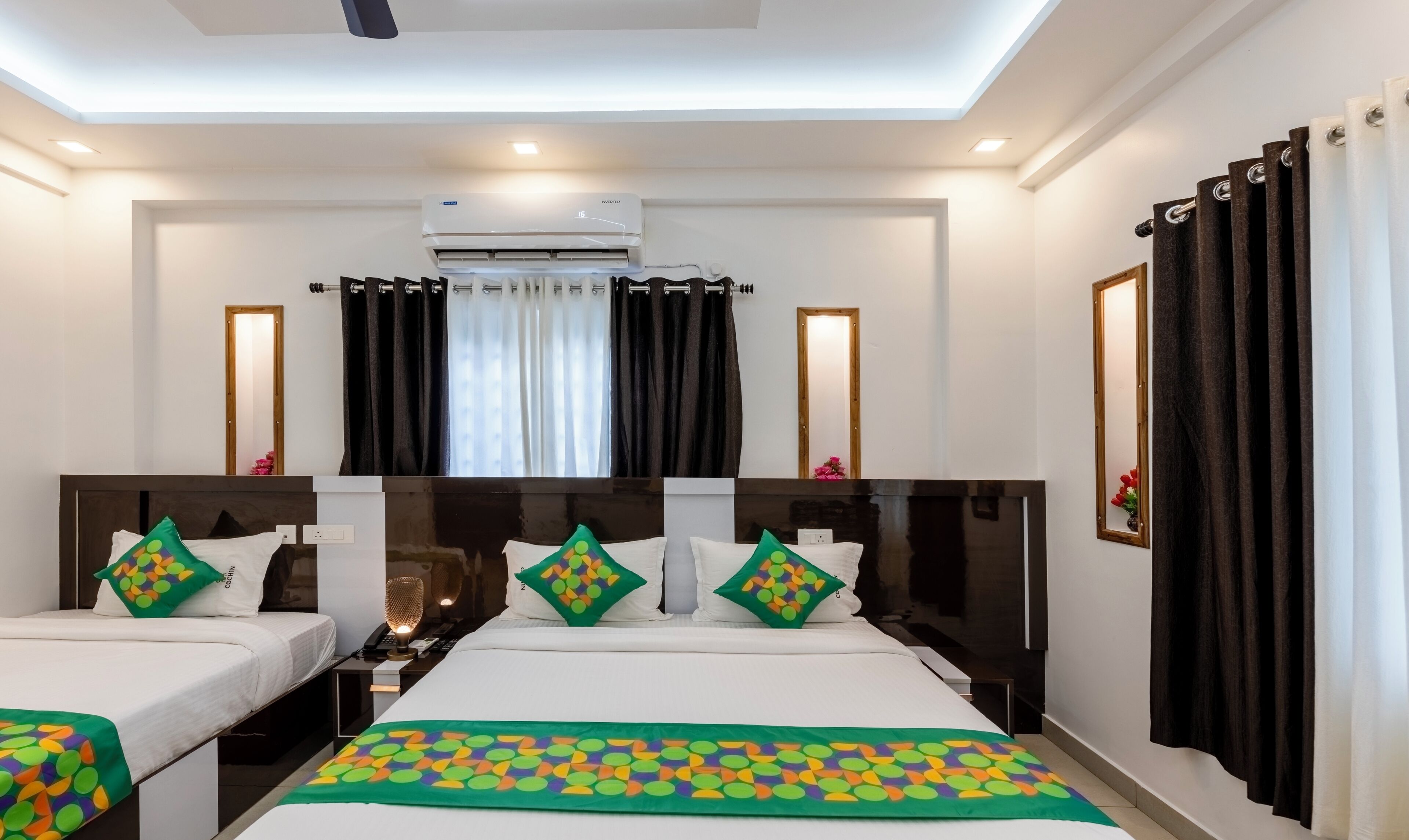 Une chambre d’hôtel avec deux lits, un ventilateur de plafond, un appareil de climatisation, une table de chevet avec une lampe et une fenêtre avec des rideaux.