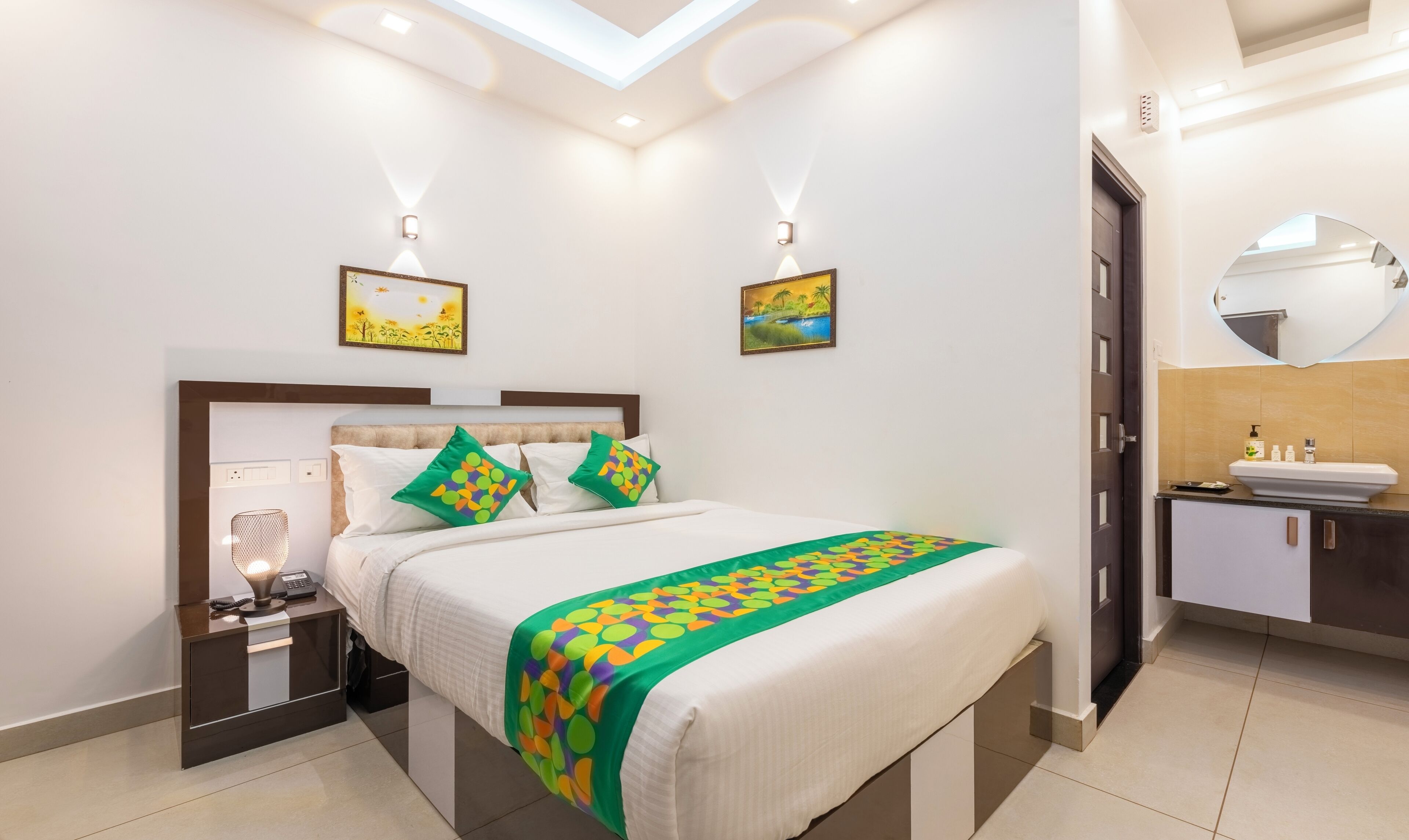 A modern hotel room with a neatly made bed, a bedside table with a lamp and phone, a bathroom with a sink and mirror, and two wall-mounted light fixtures.
