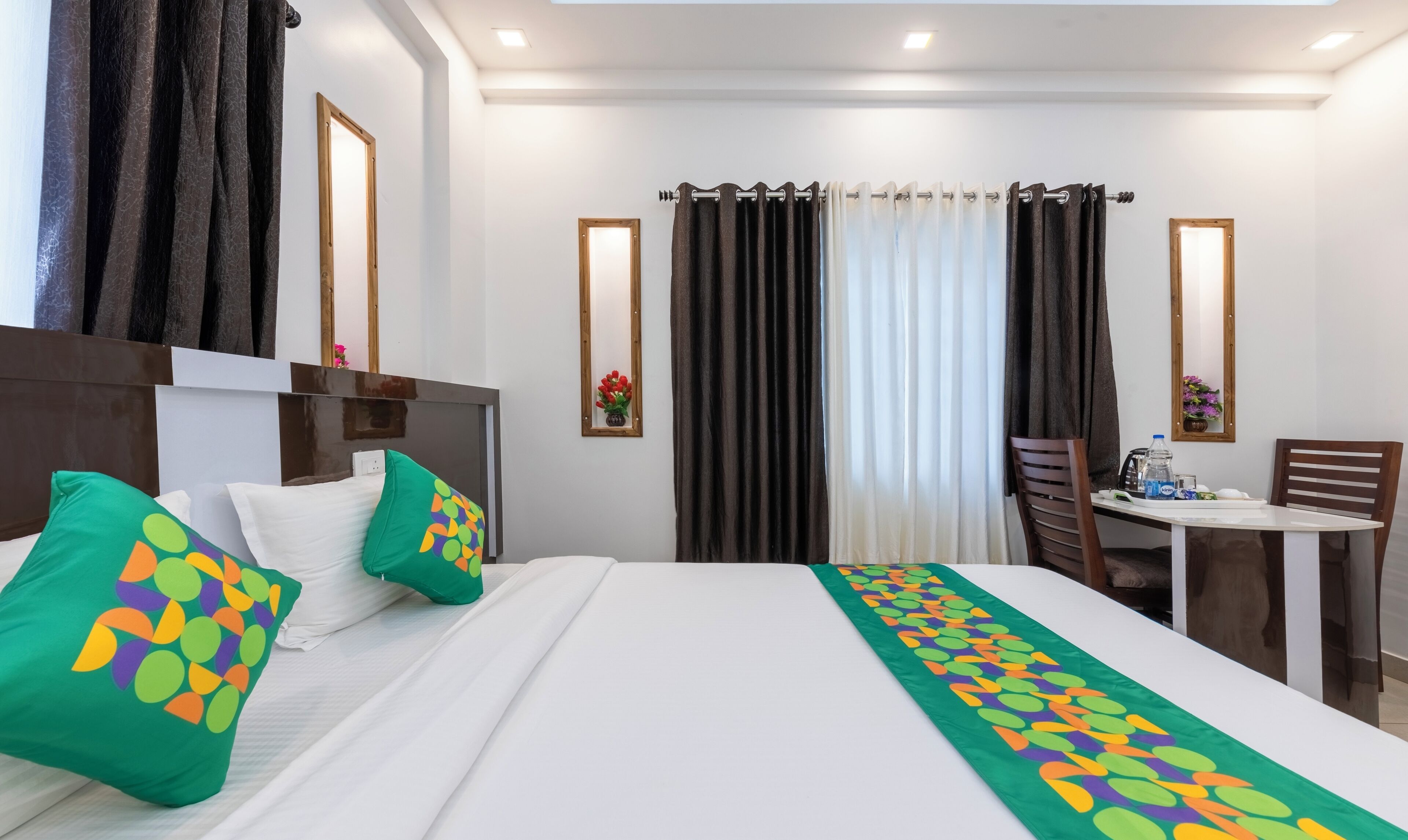 Une chambre d’hôtel avec un lit, un bureau et une chaise. Il y a des rideaux aux fenêtres et un miroir au mur.