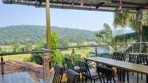An outdoor seating area with tables and chairs, a view of a green landscape, and a metal roof structure. - Big House Inn  (Calangute)
