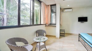 A room with two wicker chairs, a small glass table, a bed with a blue mattress, a TV mounted on the wall, and an air conditioning unit. - Big House Inn  (Calangute)