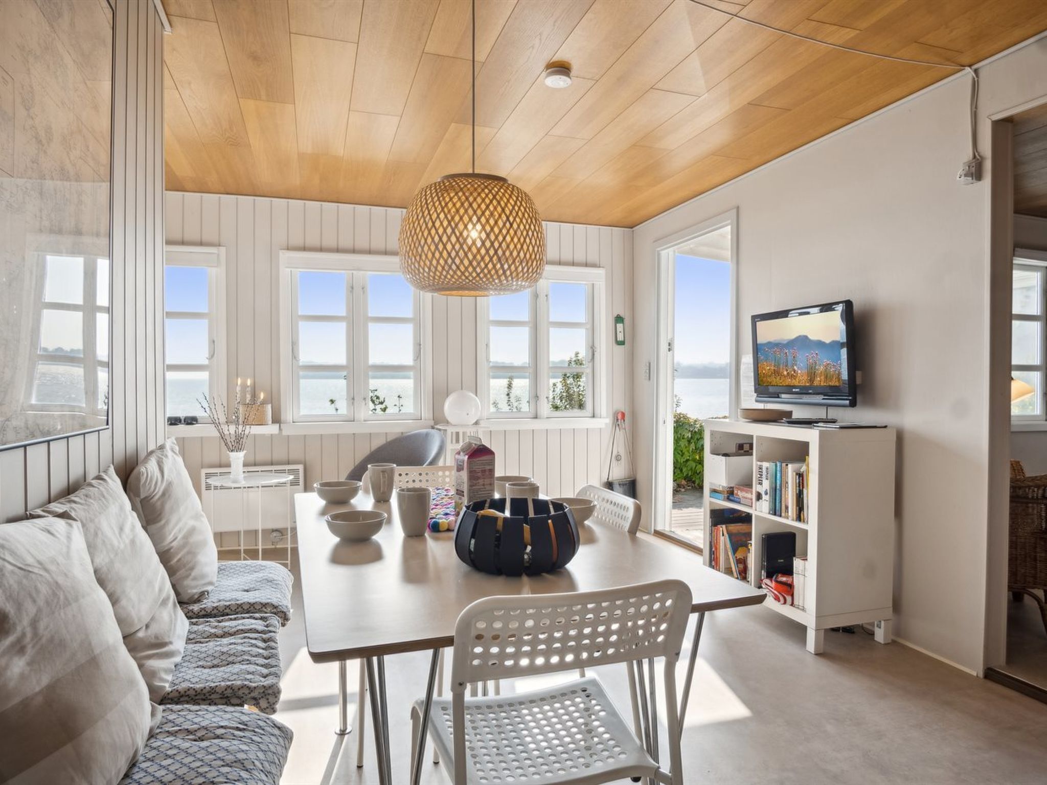 A modern living room with a wooden ceiling, white walls, and large windows. There is a dining table with chairs, a sofa, and a television on a stand.