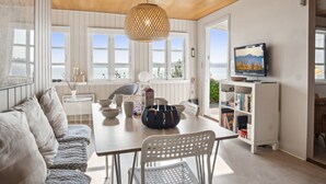 A modern living room with a wooden ceiling, white walls, and large windows. There is a dining table with chairs, a sofa, and a television on a stand. - "Drifa" - 20m from the sea by Interhome (Hejls)