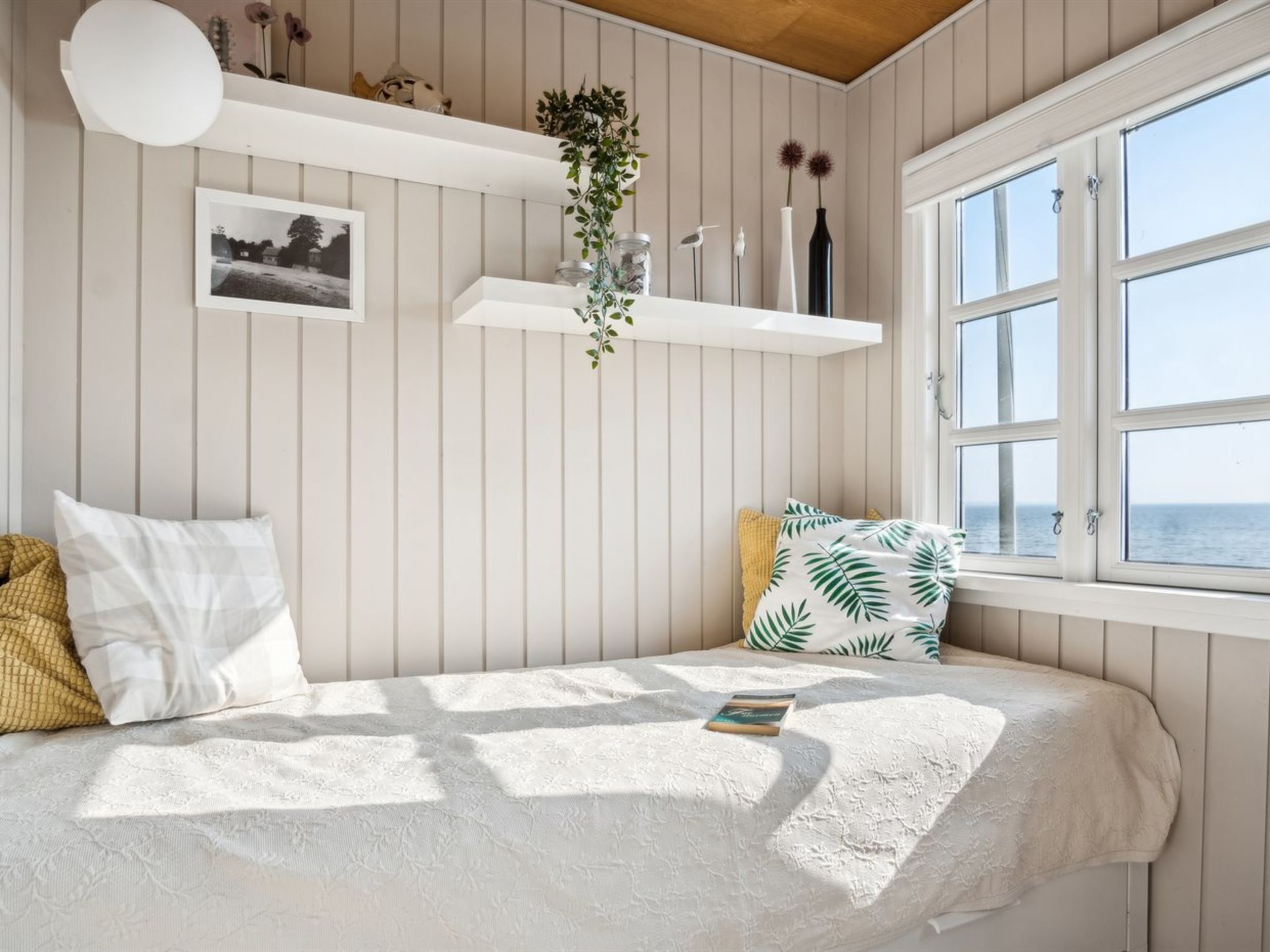 Une chambre à coucher avec un lit, une fenêtre donnant sur un beau paysage, une plante suspendue et une étagère ornée d’objets décoratifs.