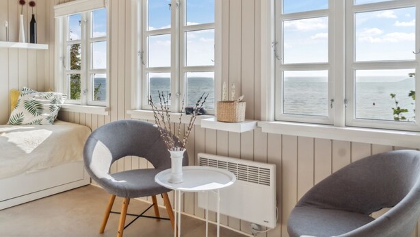 A room with a bed, two grey chairs, a small white table, and a view of the sea through large windows. - "Drifa" - 20m from the sea by Interhome (Hejls)