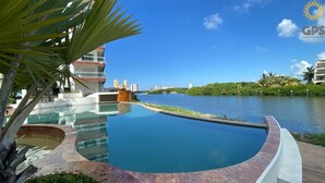 Une piscine à bord incurvé, entourée d’un espace paysager agrémenté de palmiers et offrant une vue sur les gratte-ciel d’une ville, visibles de l’autre côté de l’étendue d’eau.